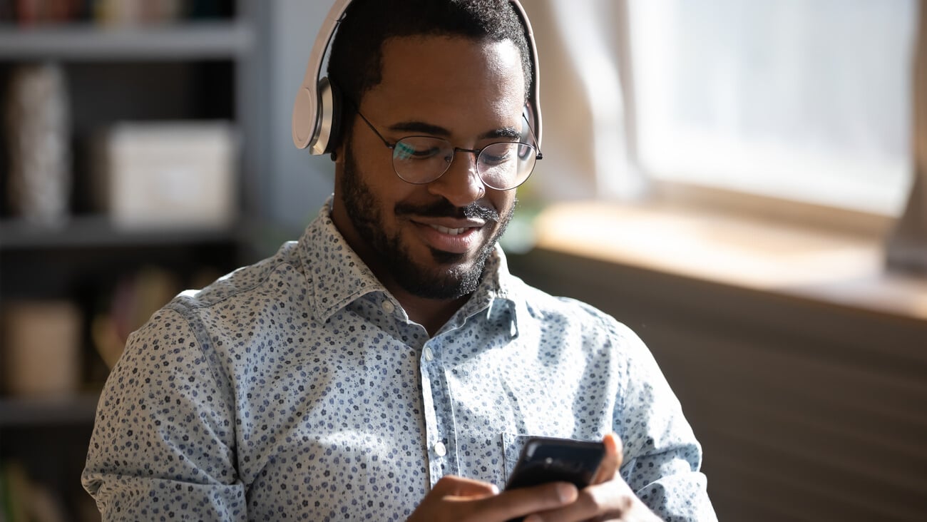African man wear headphones holds cellphone choose track listens digital music or podcast, using video streaming service. Language learning platform most downloaded education app advertisement concept; Shutterstock ID 1755781163