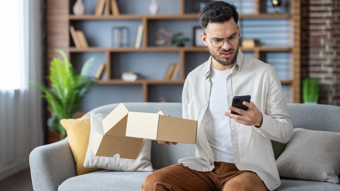 Angry young Indian man sitting on sofa at home, holding open parcel box and looking disappointedly at mobile phone, dialing delivery service with complaints.; Shutterstock ID 2480562869
