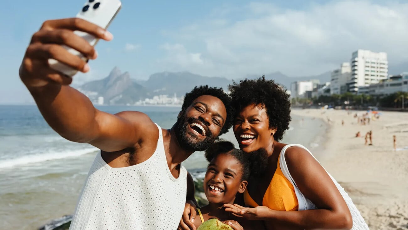 A cheerful Brazilian family enjoys a sunny day at a beach in Rio de Janeiro, capturing the moment with a selfie. The scene showcases joy and togetherness, with the iconic cityscape in the background.; Shutterstock ID 2545766889