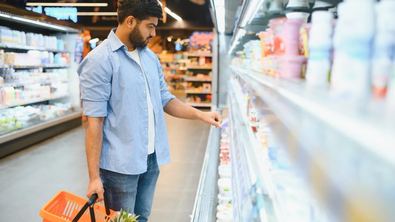 Concentrated young indian man choosing dairy product from refrigerator shelf, holding shopping basket with pineapple, shopping in supermarket; Shutterstock ID 2669141919