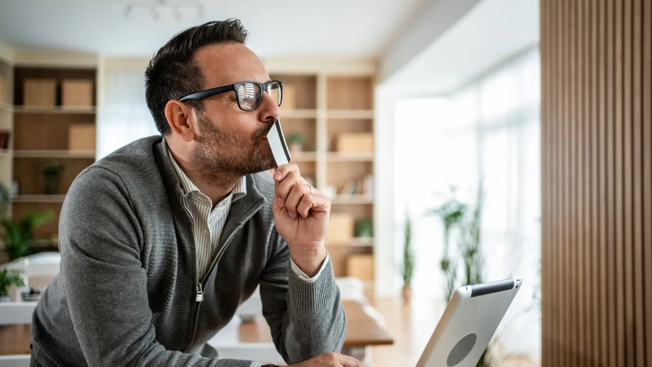Man holding a credit card to his face while thoughtfully using a tablet at home, contemplating online purchases, budgeting and digital payments in a relaxed living room setting; Shutterstock ID 2704848009