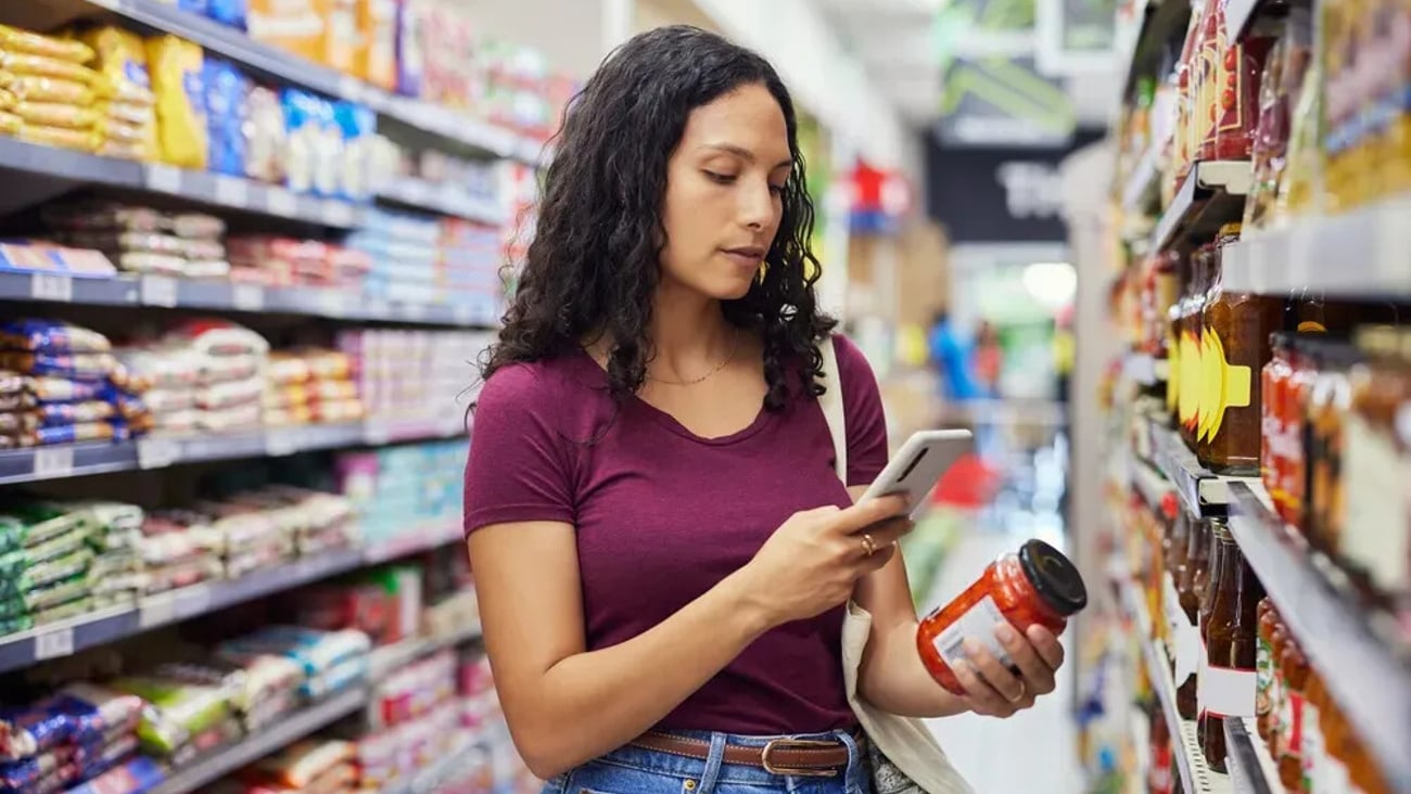 Multiethnic young woman checking product label while holding a smartphone in grocery aisle. Focused shopper using phone while scanning nutrition label of food product. Casual customer standing in supermarket with shopping bag and using mobile phone to check the quality.; Shutterstock ID 2734098113