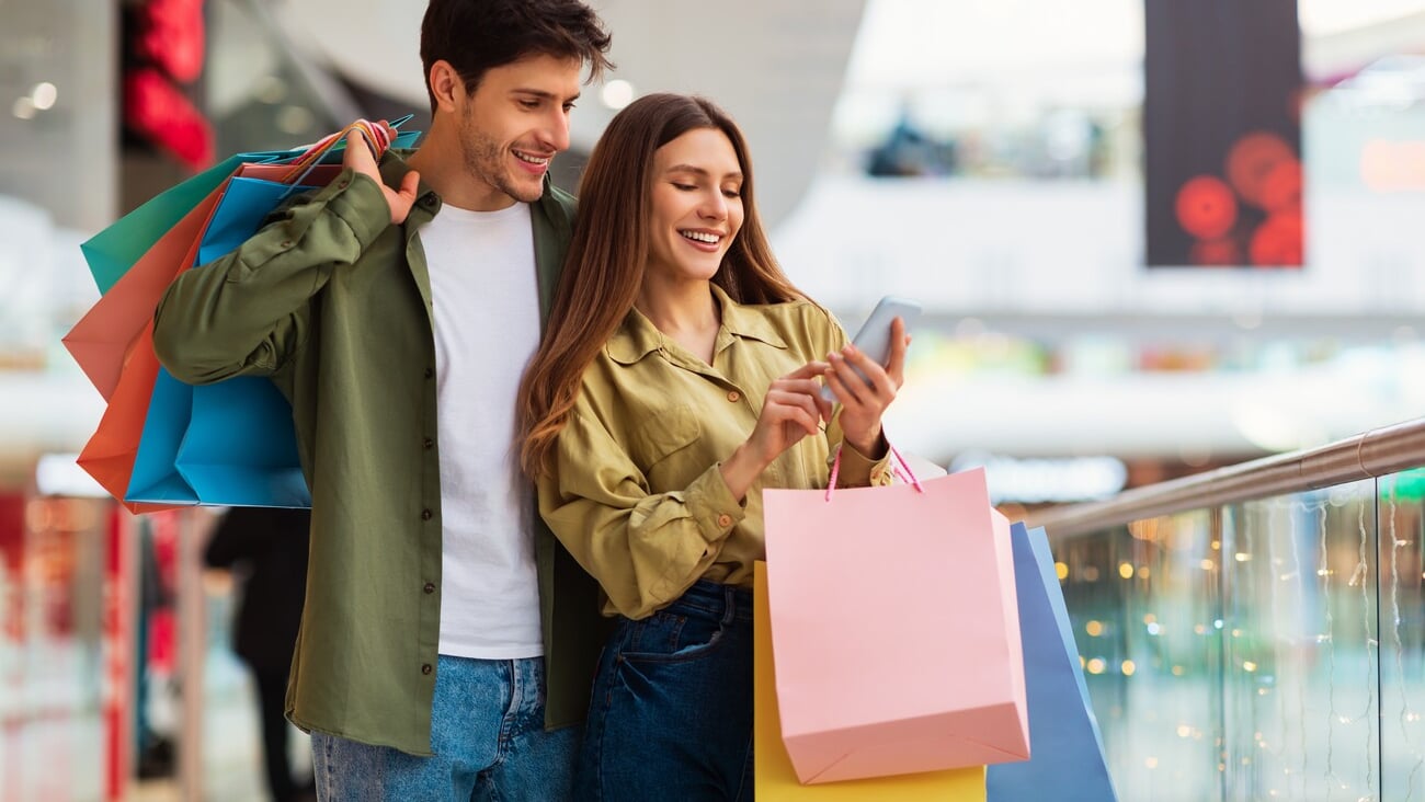 Buyers Couple Shopping Using Cellphone Holding Colorful Shopper Bags Standing In Mall. Happy Customers Using Application Purchasing Clothes Online Via Smartphone. Ecommerce And Shopaholism; Shutterstock ID 2116844222