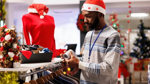 Cheerful man employee working in store during Christmas season discounts, arranging fashion items in festive decorated shop. African american mall assistant preparing clothing for shopping frenzy.; Shutterstock ID 2402348579