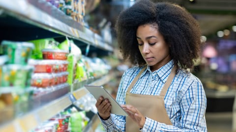 A grocery store employee in an apron checks the inventory on the shelves using a tablet. The worker appears focused and attentive, ensuring the store is well-stocked and organized for customers.; Shutterstock ID 2483544541