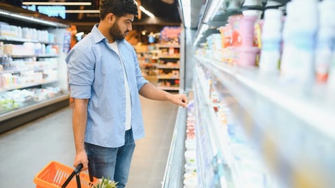 Concentrated young indian man choosing dairy product from refrigerator shelf, holding shopping basket with pineapple, shopping in supermarket; Shutterstock ID 2669141919