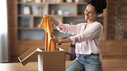 Satisfied customer attractive young woman opening paper box, holding stylish brand new clothes and smiling at camera, female buyer checking her delivery at home, copy space; Shutterstock ID 2201003471