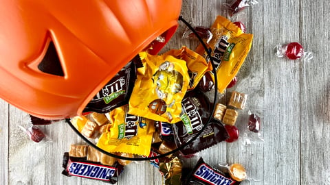 Orange plastic pumpkin bucket with spilled Halloween candy on a wood surface; Shutterstock ID 1179428302