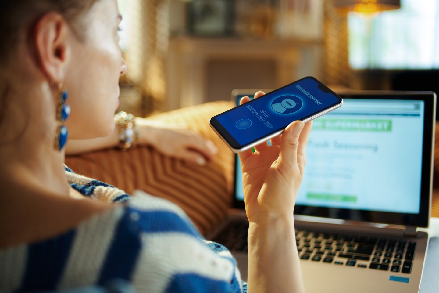 Over shoulder shot of young woman laying on sofa at modern home in sunnny hot summer day getting help from voice robot support while order food digital at online supermarket.; Shutterstock ID 1444176149