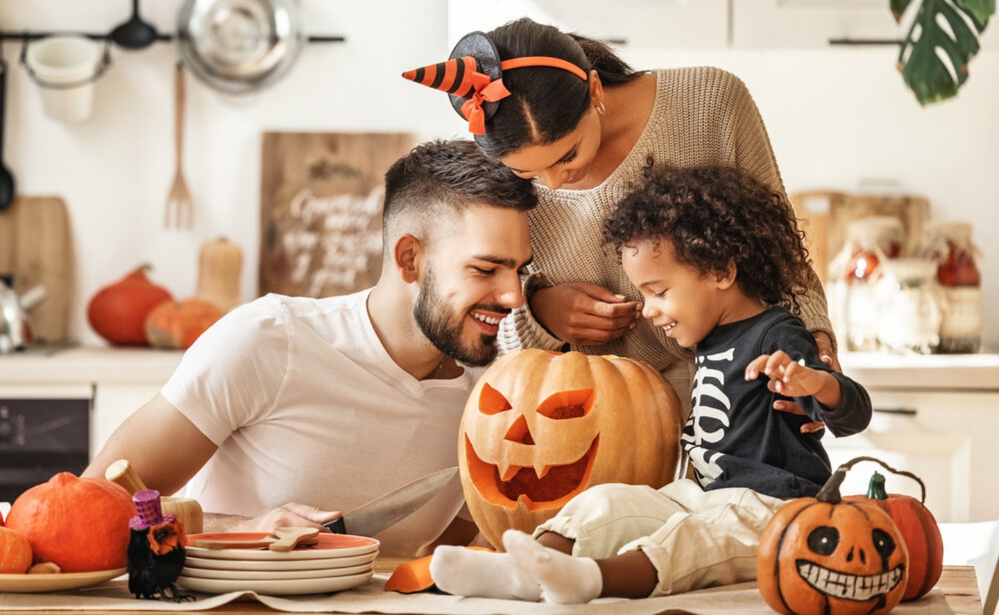 Cheerful multi ethnic family parents with son smiling  while creating jack o lantern from pumpkin during Halloween celebration in kitchen at home.; Shutterstock ID 1833746497