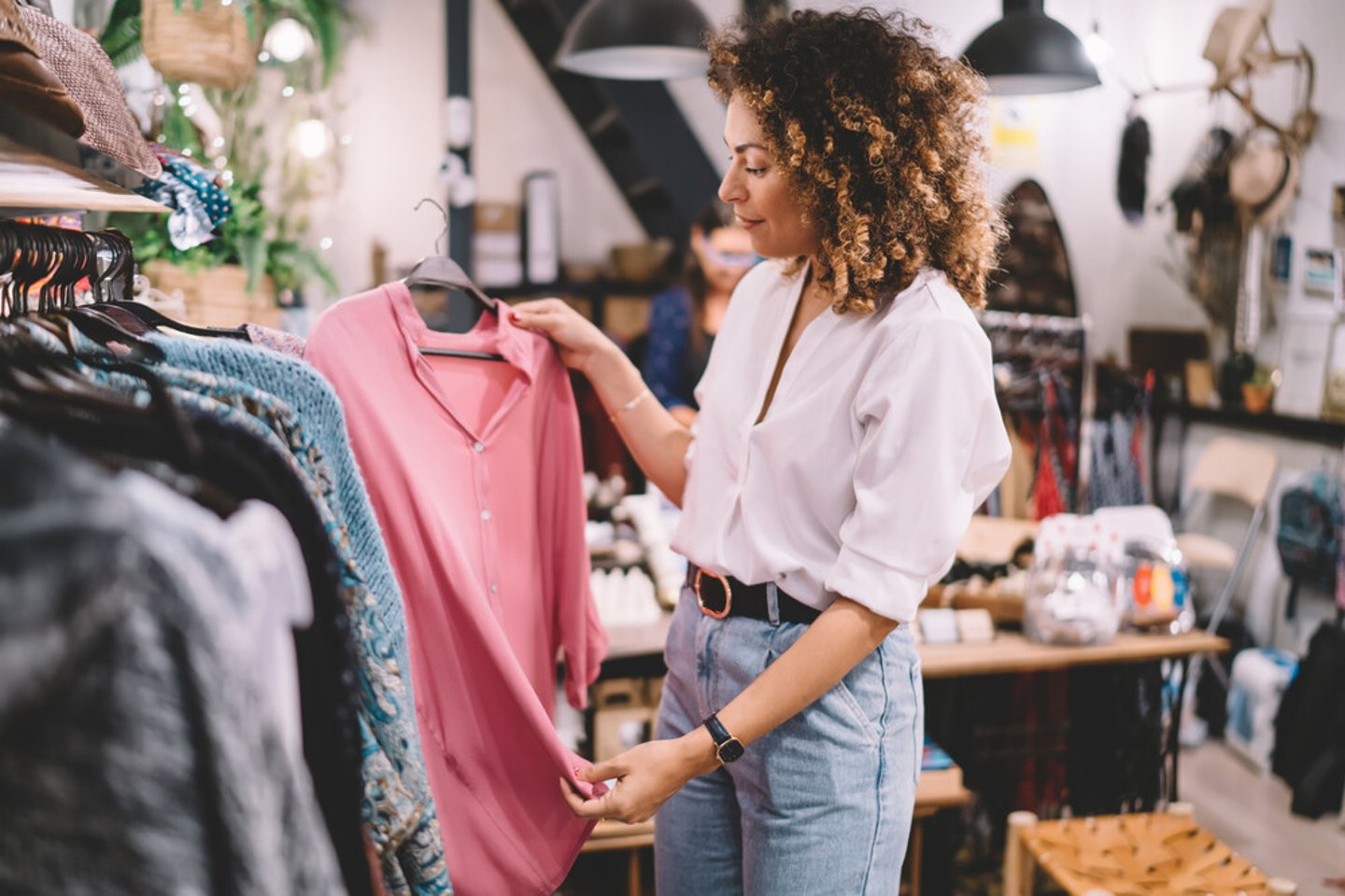 Caucaisan female customer with curly hair buying clothes in boutique using fitting concept while choosing, professional stylist creating trendy look concentrated on selection in designer shop; Shutterstock ID 1854949711