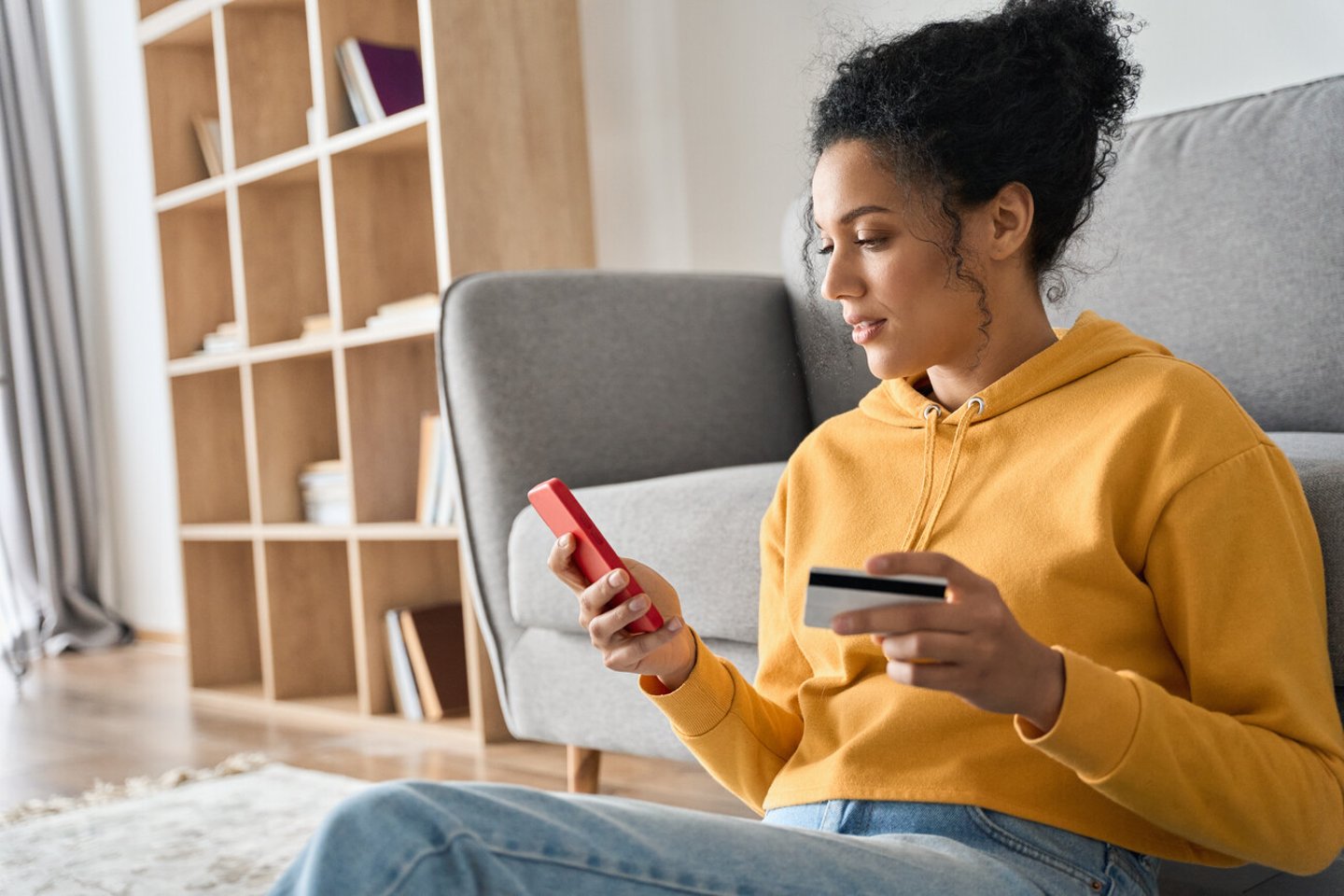 Young adult African American female consumer holding credit card and smartphone sitting on floor at home doing online banking transaction. E commerce virtual shopping, secure mobile banking concept.; Shutterstock ID 1979496134