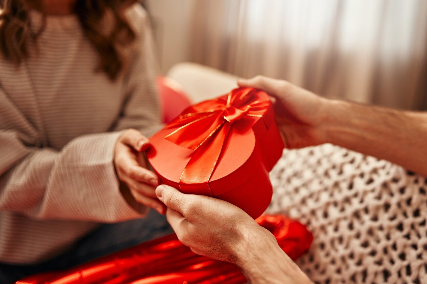 Happy Valentine's Day. Young couple in love exchanging gifts while sitting on the sofa in the living room at home. A man giving a heart-shaped gift box to his beloved woman.; Shutterstock ID 2409568521