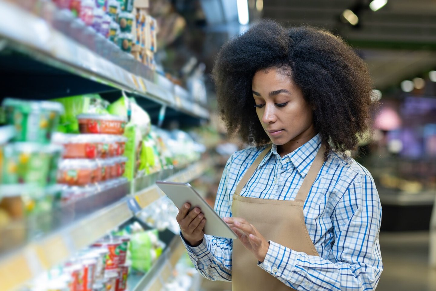 A grocery store employee in an apron checks the inventory on the shelves using a tablet. The worker appears focused and attentive, ensuring the store is well-stocked and organized for customers.; Shutterstock ID 2483544541