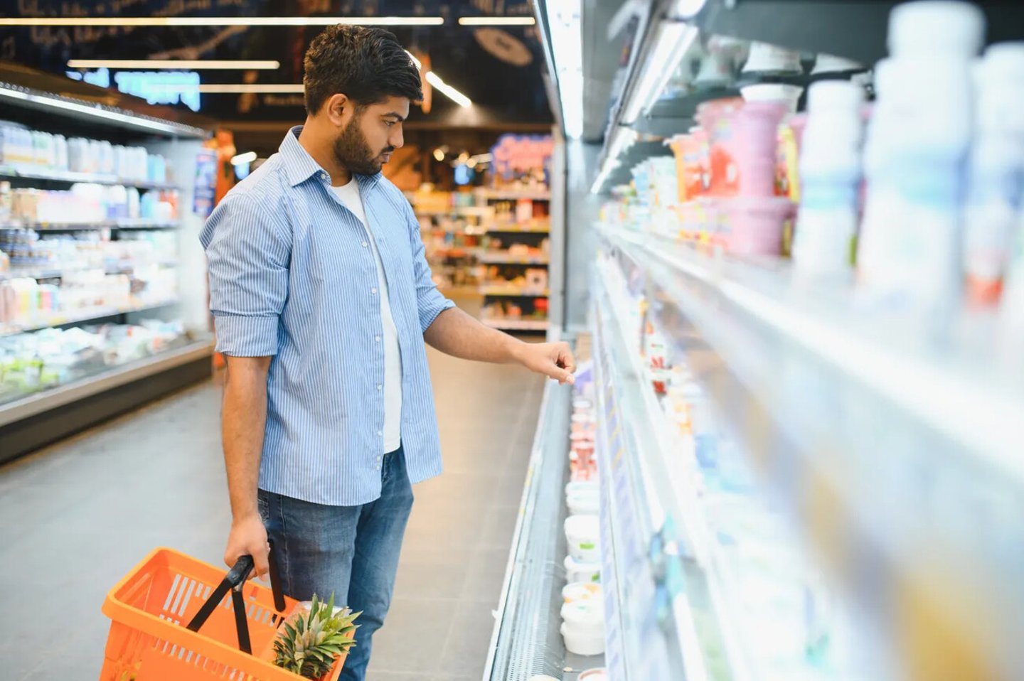 Concentrated young indian man choosing dairy product from refrigerator shelf, holding shopping basket with pineapple, shopping in supermarket; Shutterstock ID 2669141919