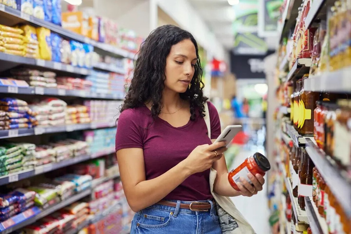 Multiethnic young woman checking product label while holding a smartphone in grocery aisle. Focused shopper using phone while scanning nutrition label of food product. Casual customer standing in supermarket with shopping bag and using mobile phone to check the quality.; Shutterstock ID 2734098113