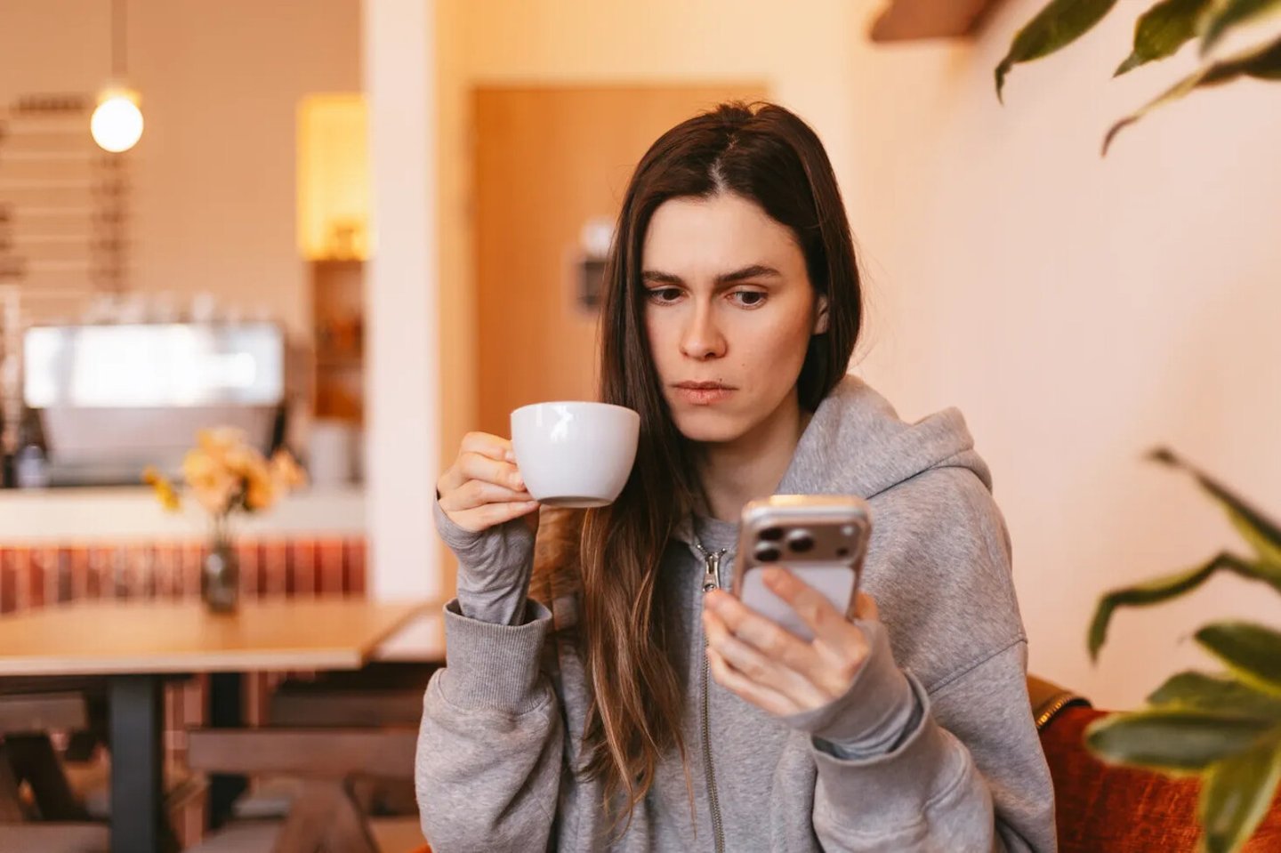 Serious young woman in grey hoodie holding white cup of coffee and looking at smartphone screen with concerned face expression sitting in cozy cafe interior during morning break time; Shutterstock ID 2757270449