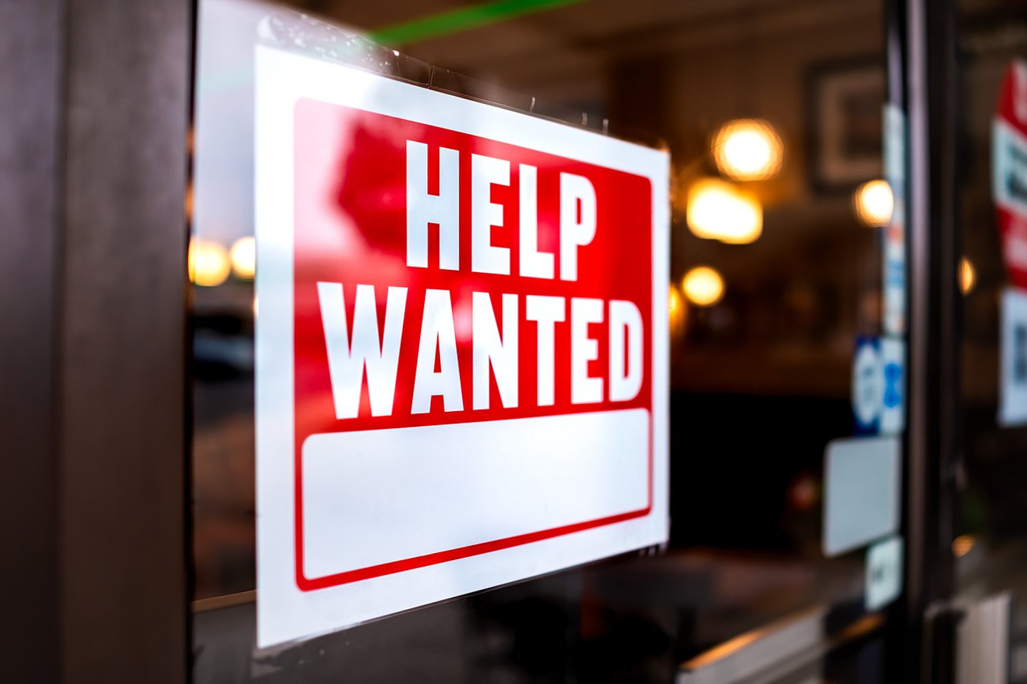 Sign text closeup for help wanted with red and white colors by entrance to store shop business building during corona virus covid 19 pandemic; Shutterstock ID 1840085851