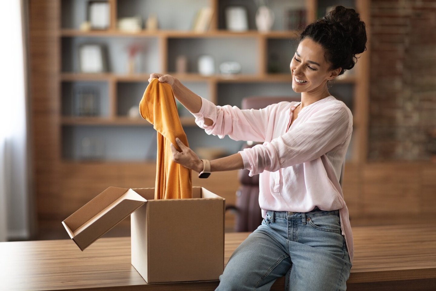 Satisfied customer attractive young woman opening paper box, holding stylish brand new clothes and smiling at camera, female buyer checking her delivery at home, copy space; Shutterstock ID 2201003471