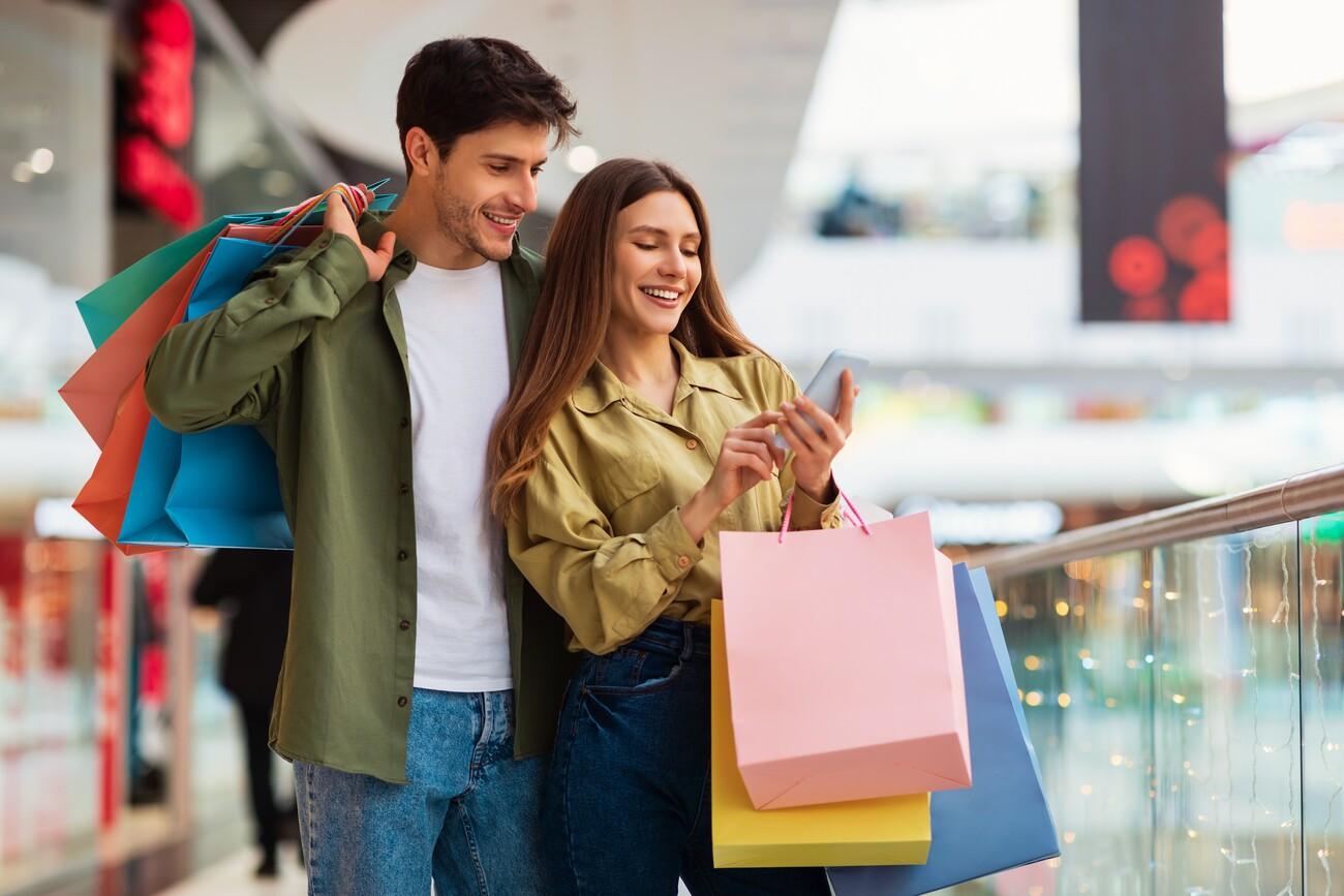 Buyers Couple Shopping Using Cellphone Holding Colorful Shopper Bags Standing In Mall. Happy Customers Using Application Purchasing Clothes Online Via Smartphone. Ecommerce And Shopaholism; Shutterstock ID 2116844222