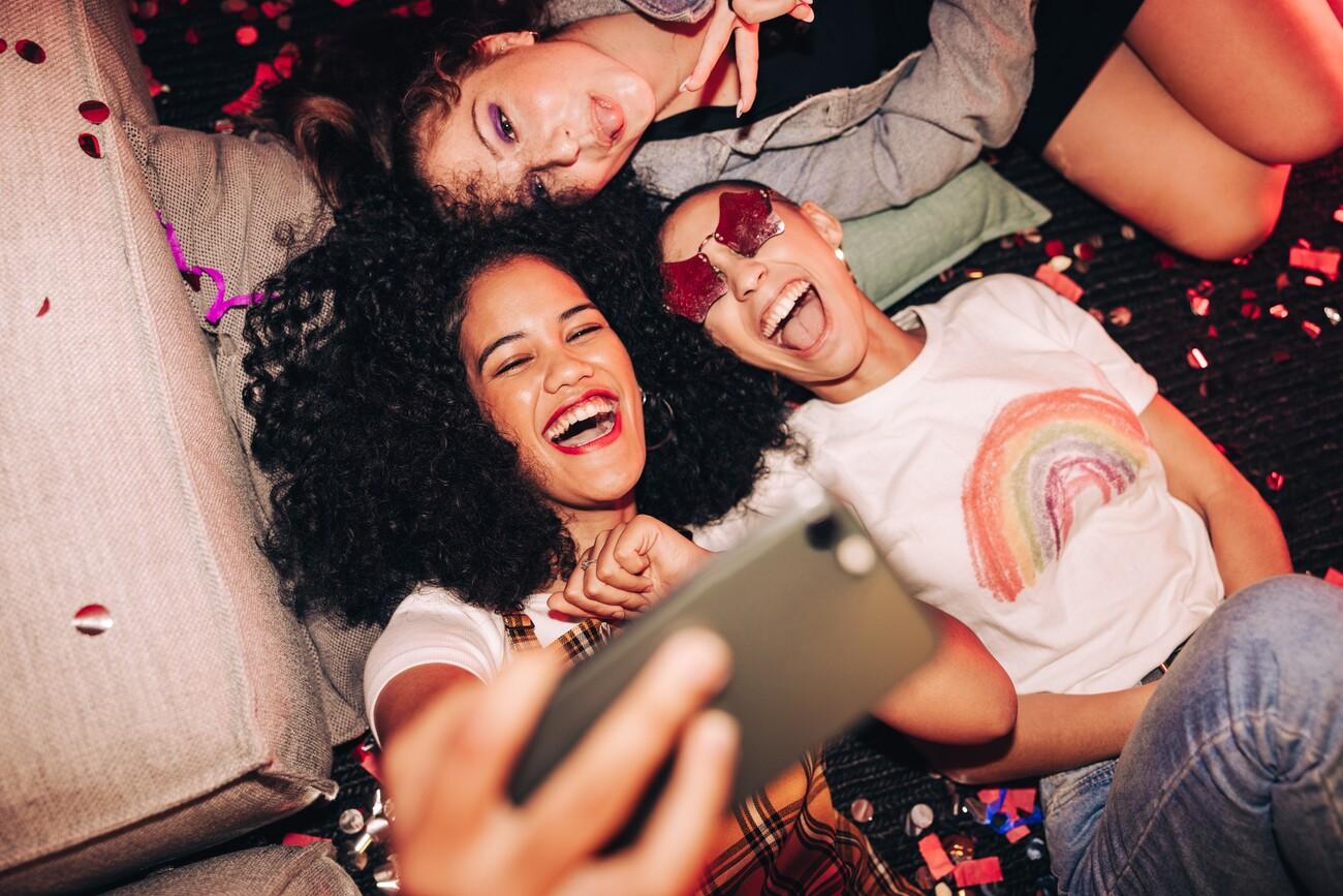 Vibrant selfies for vibrant people. Overhead view of three happy friends taking a selfie while lying on the floor at a house party. Group of cheerful female friends having fun together on the weekend.; Shutterstock ID 2135759921