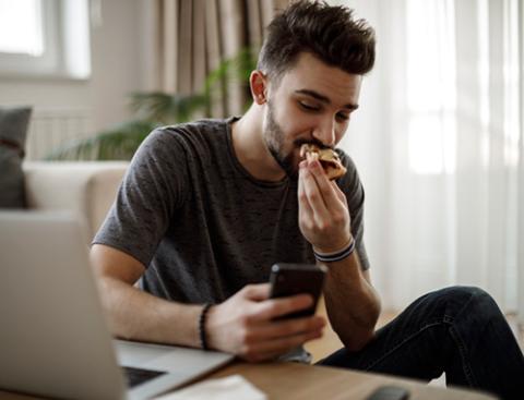 man eating pizza near laptop using mobile phone